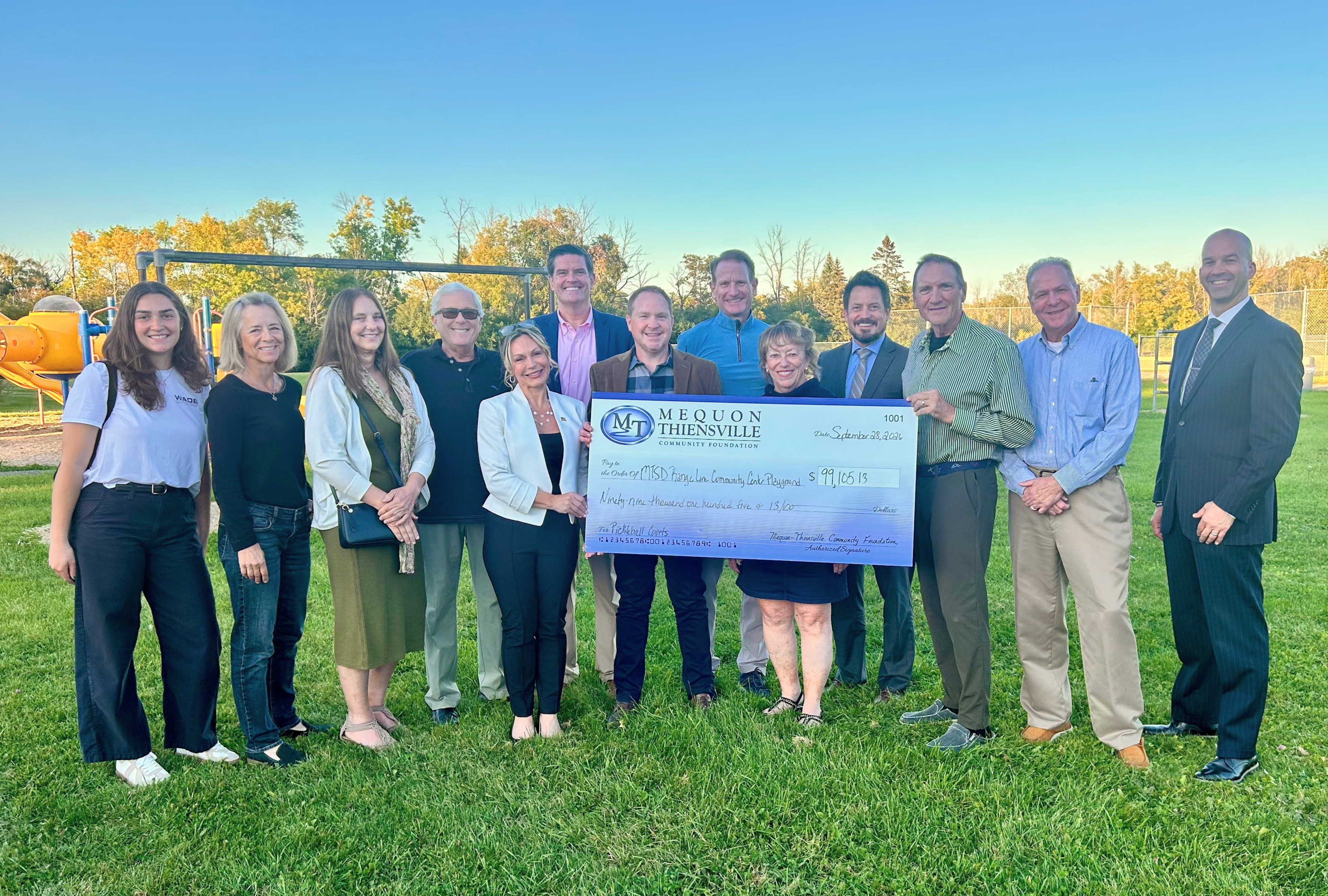 A group of people standing outdoors on a grassy field, holding a large ceremonial check together.